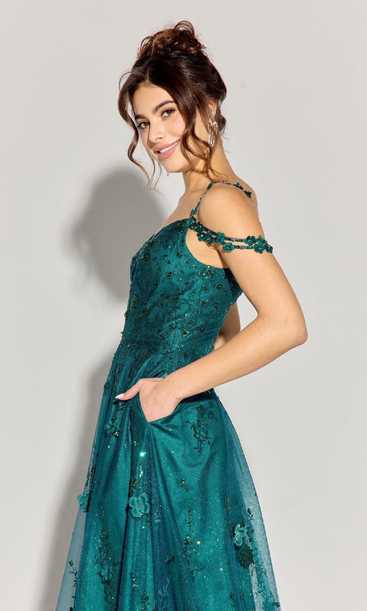 A woman stands sideways, hand in pocket, wearing the Eureka 9980 Long Glitter-Tulle A-Line Prom Dress with floral embellishments against a plain light background.